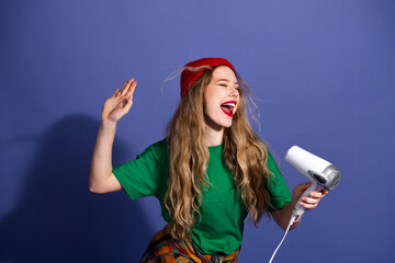 Young woman with red beanie sings and uses a hair dryer in a casual studio scene wearing a green shirt plaid waist wrap and a bold blue background