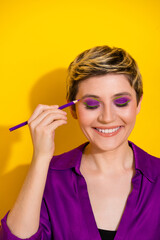 Obraz premium Young woman applies purple eye makeup against a bright yellow backdrop wearing a purple blouse showcasing glamour