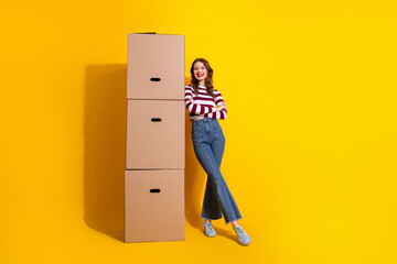 Young woman with striped top stands leaning on a tall stack of cardboard boxes against a bright yellow background in a casual stylish lifestyle moment