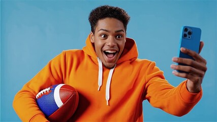 Excited Young Male Sports Fan in an Orange Hoodie Taking a Selfie or Recording a Reaction Video with his Smartphone and Holding an American Football Against a Blue Background - Powered by Adobe