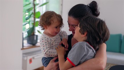 Mother holding toddler and older child together near window, children gently touching hands while sharing soft smile in peaceful family moment - Powered by Adobe