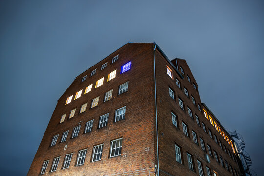 A low-angle shot captures a historical red brick industrial building illuminated against a dark evening sky, featuring numerous windows with contrasting warm and blue interior light.