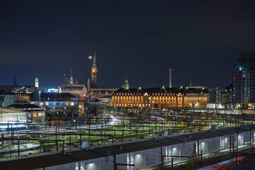 Fototapeta premium A panoramic, long-exposure night shot captures a vibrant urban Copenhagen skyline featuring historic architecture and the complex rail infrastructure of a metropolitan city.
