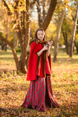 A young woman stands in an autumn forest. She's wearing a long red plaid dress and a warm cape, a romantic, vintage style. She's holding a pistol.