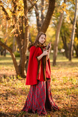 A young woman stands in an autumn forest. She's wearing a long red plaid dress and a warm cape, a romantic, vintage style. She's holding a pistol.