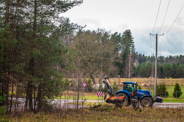 Blue tractor with front-mounted mower clearing vegetation along a rural roadside near fields, forest, and power lines. © Valters