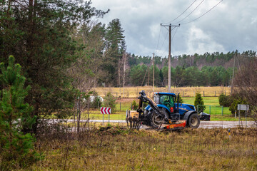 Blue tractor with front-mounted mower clearing vegetation along a rural roadside near fields, forest, and power lines. © Valters