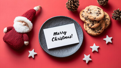 Fictional Christmas decoration with Santa Claus toy on a red table with a "Merry Christmas" note written in black on white paper placed onto a grey plate. Decorations: white stars cookies, pine cones.