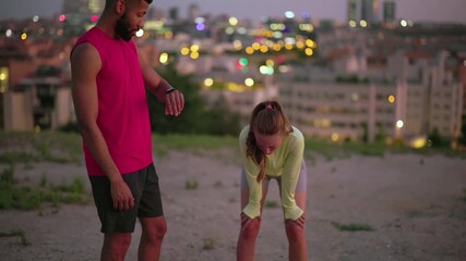 Multiracial athletic couple taking a break after running in the city at dusk. Young exhausted woman is catching her breath while the man is checking their performance on his fitness tracker - Powered by Adobe