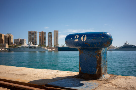 Old blue mooring bollard with number twenty painted in white at the port, with blurred city buildings and yachts on calm turquoise sea under clear sunny sky