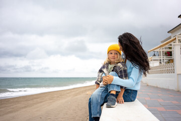 Affectionate mother holding her young son in a yellow beanie while sitting on a seaside wall, sharing a warm moment together against the backdrop of the beach and cloudy sky
