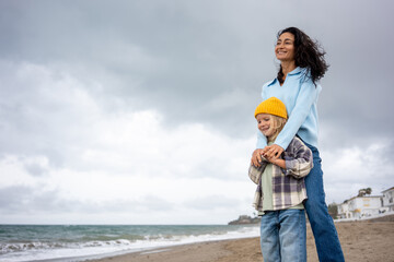Happy mother embraces her young son on the beach, woman in blue sweater and child in yellow beanie smiling together by the ocean on a cloudy day