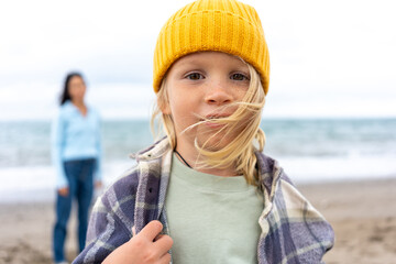 Close-up portrait of a blond child in yellow beanie and plaid jacket, standing on a windy beach with hair blowing, while a woman in blue sweater appears blurred in the background