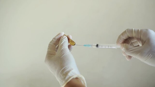 Gloved hands drawing liquid medication from an open glass ampoule using a syringe. Sterile medical procedure suitable for training, treatment, or pharmaceutical visuals.
