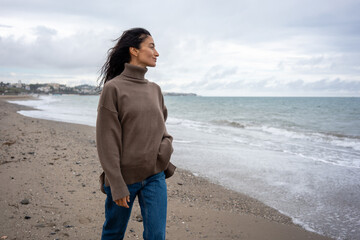 Stylish woman walking along sandy beach near ocean waves, wearing brown sweater, jeans, and sneakers, with wind blowing through her hair on a cloudy day