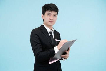 A cheerful and diligent young Asian businessman in a black suit, smiling while writing or checking a document on a clipboard against a light blue studio background