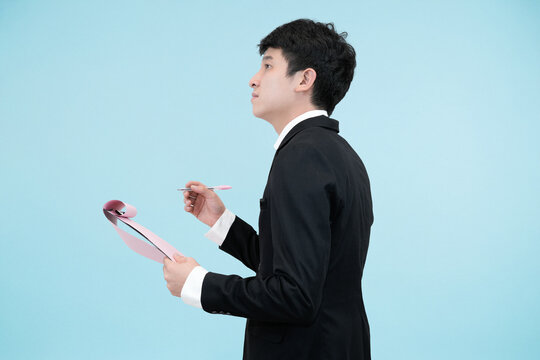 A serious young Asian businessman in a black suit, writing or marking a document on a clipboard with a demandin expression in studio blue background - Powered by Adobe