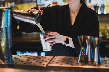 A female bartender in a black shirt skillfully pours a green cocktail from a metal shaker into a glass in a dark, moody bar setting