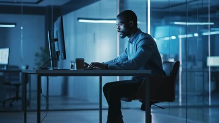 A man sits at a desk in a bluelit office focused on a computer amid glass walls, bathed in blue glow - Powered by Adobe