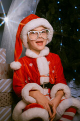 Child in a Santa costume on a sleigh by a festive tree with glowing blue lights, surrounded by wrapped presents