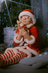 Cute boy dressed as Santa Claus hugging a soft reindeer plush toy, enjoying the holiday spirit by a Christmas tree with gifts.