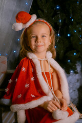 Girl dressed as a red elf with a soft cape sitting on a sled near a Christmas tree decorated with blue lights.