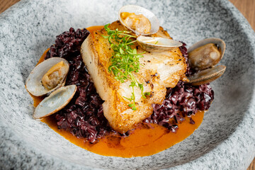 A beautifully plated dish showcasing a piece of fish on black rice with clams and garnishes