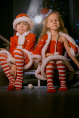 Joyful brother and sister in red and white Santa outfits embracing on a sled inside a cozy home with Christmas decorations.