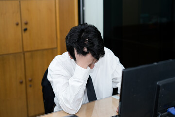 A stressed Asian office worker or businessman sitting at his desk, burying his face in his hands,...