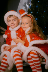 Brother and sister in Santa costumes sitting on a sled indoors, hugging joyfully near a beautifully decorated Christmas tree with sparkling lights and festive gifts.