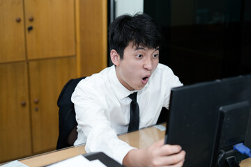 Fototapeta premium A young Asian businessman or office worker sitting at his desk, staring at the computer screen with a wide-eyed expression of shock and surprise
