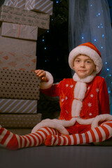 Boy in Santa costume and hat sitting by a Christmas tree with blue lights, surrounded by many wrapped gifts indoors