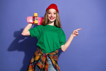 Young girl with green shirt red beanie and skateboard smiles at camera against purple background in casual streetwear fashion shoot