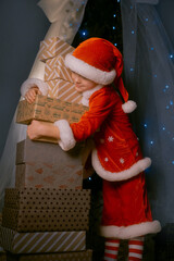Boy in Santa costume and hat standing by a Christmas tree with blue lights, hugging a stack of wrapped gifts and laughing