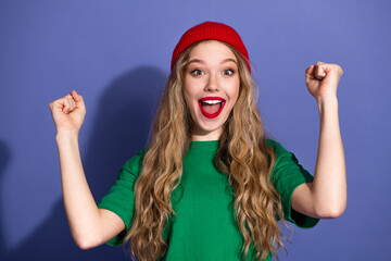 Cheerful young woman in green shirt red beanie celebrates success with raised fists against purple...