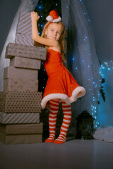 Young girl in a red Santa dress standing gracefully, leaning on a tall stack of beautifully wrapped Christmas presents.