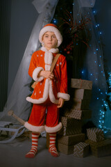 Young boy in a red and white elf costume standing by a Christmas tree decorated with holiday lights, holding a festive backpack.