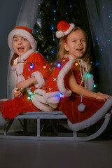 Festive New Year scene with siblings dressed as elves on a sled, completely entwined in glowing Christmas lights by a decorated tree.