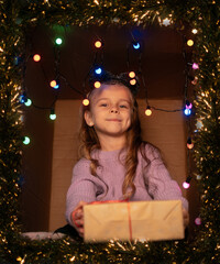 Laughing girl in a purple sweater holding her gift while sitting in a decorated box with lights and tinsel