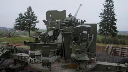 green antiaircraft gun mounted on concrete platform with pine backdrop, overcast sky and weathered paint; close view of turret mechanism, breech and elevation gears, museum display feel,