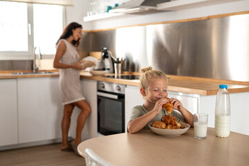 Cute child eating fresh croissant with glass of milk at kitchen table while mother preparing breakfast in background, family morning routine at home