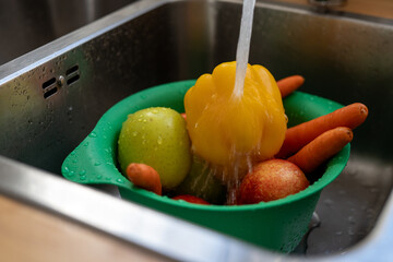 Yellow bell pepper, green apple, red nectarine, and carrots being washed under running water in green colander inside kitchen sink