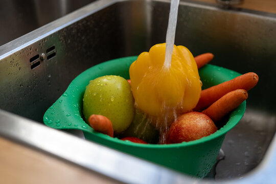 Yellow bell pepper, green apple, red nectarine, and carrots being washed under running water in green colander inside kitchen sink