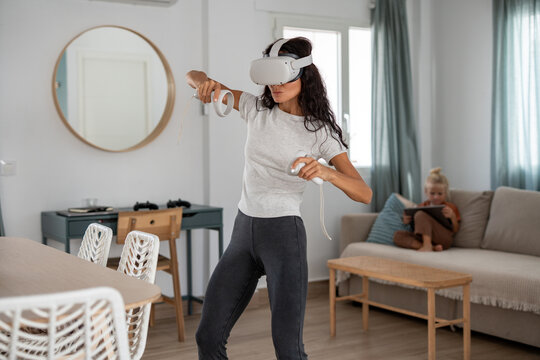 Focused woman wearing VR headset and playing virtual reality game with controllers while child uses digital tablet on sofa in modern living room