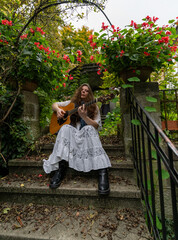 Young woman musician playing acoustic guitar on stone garden steps surrounded by vibrant red flowers and green foliage