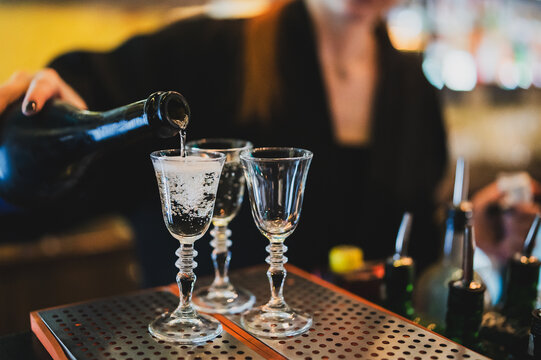 Bartender pouring clear alcohol into elegant shot glasses on a metal bar surface. Close-up shot, vibrant bar atmosphere, preparing drinks, nightlife, service - Powered by Adobe