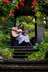 young woman playing acoustic guitar seated in lush garden archway with vibrant flowers and greenery