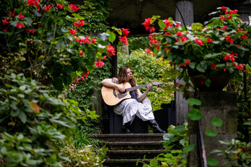 Young woman playing acoustic guitar outdoors in lush green garden with red flowers around