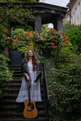 Musician woman standing on stone garden stairs holding acoustic guitar with lush greenery and red flowers around