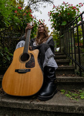 Young woman musician sitting on outdoor steps holding acoustic guitar wearing casual outfit